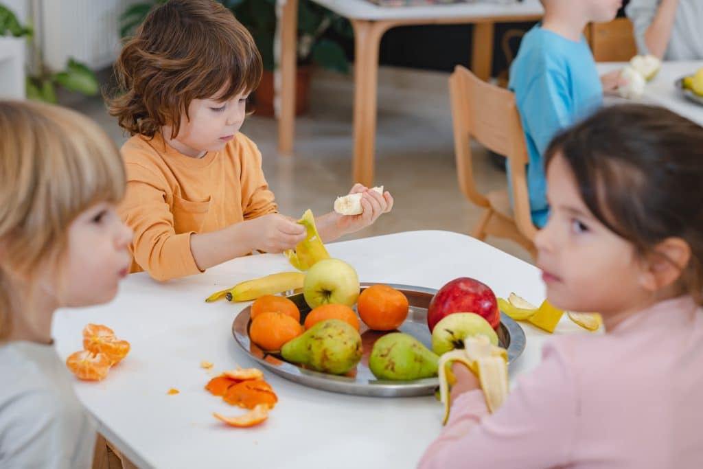 Kinderen lunchen bij de kinderopvang. Er staat een grote schaal fruit op tafel. 